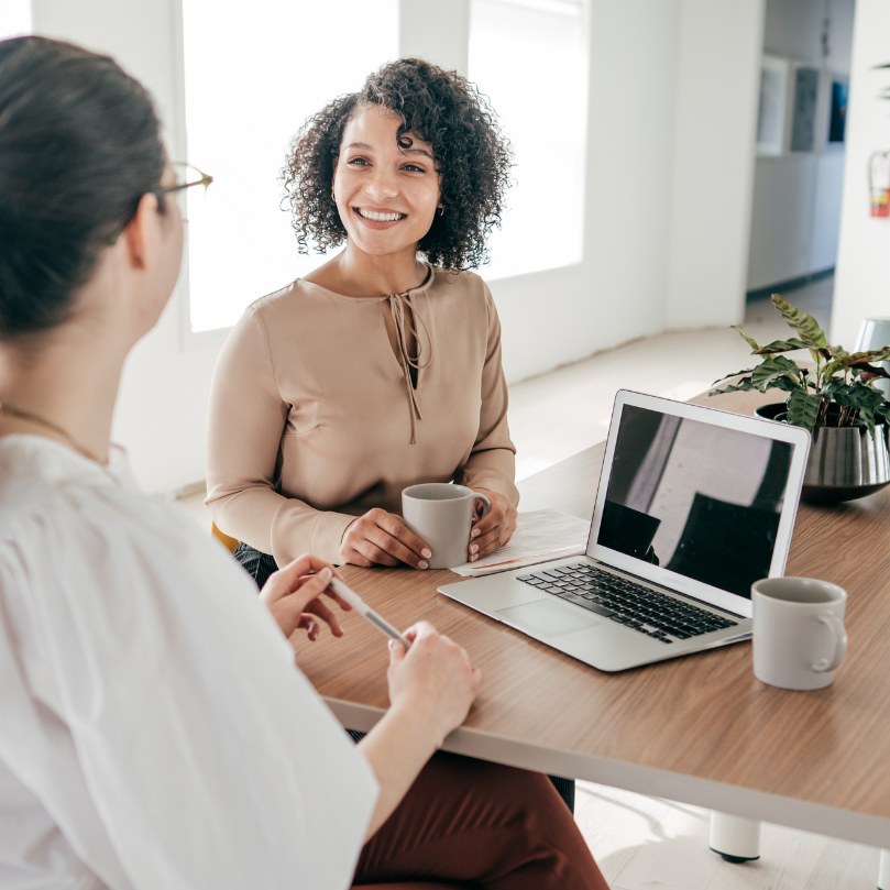 two women sitting at a table with a laptop and coffees, one smiling at the other